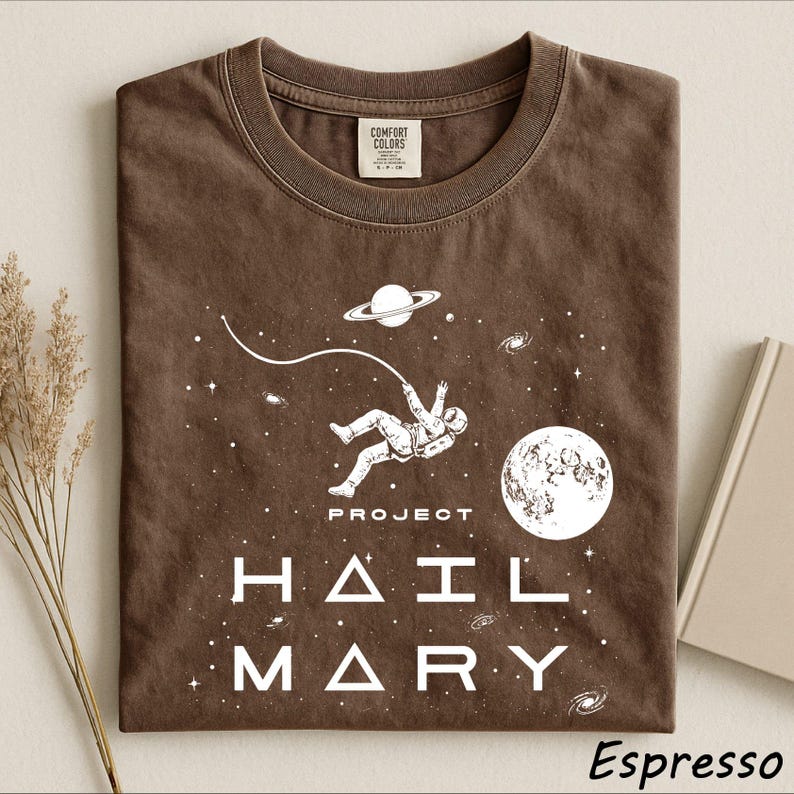 May include: A brown t-shirt featuring a white graphic of an astronaut floating in space with a planet, a moon, and stars. The text "PROJECT HAIL MARY" is printed in white below the astronaut. The shirt has a crew neckline and a Comfort Colors label.
