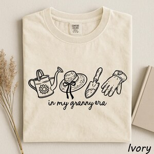 A beige t-shirt with a graphic design featuring a watering can, a hat, a shovel, and a pair of gloves, along with the text "in my granny era".