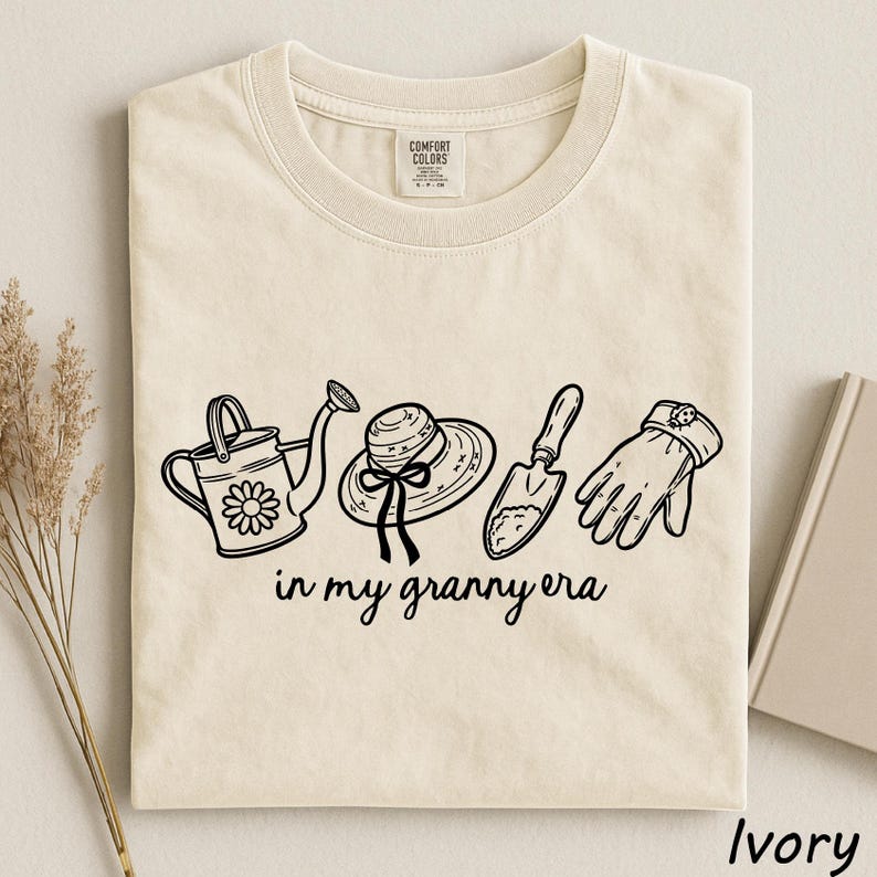 A beige t-shirt with a graphic design featuring a watering can, a hat, a shovel, and a pair of gloves, along with the text "in my granny era".