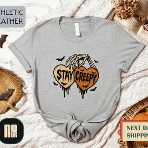 a gray t-shirt with a Halloween-themed graphic, including a skeleton hand holding a heart with the text "STAY CREEPY" and bats. The t-shirt is displayed on a white background, surrounded by various Halloween-themed items such as bats, a pumpkin, and a woven basket.