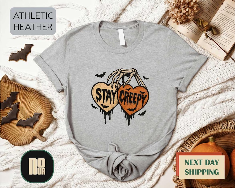 a gray t-shirt with a Halloween-themed graphic, including a skeleton hand holding a heart with the text "STAY CREEPY" and bats. The t-shirt is displayed on a white background, surrounded by various Halloween-themed items such as bats, a pumpkin, and a woven basket.