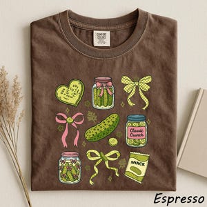 a brown t-shirt with a variety of colorful illustrations, including jars, a heart, a bow, a pickle, and a bag of chips, all set against a white background.