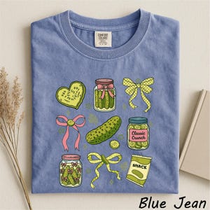 A blue t-shirt with a graphic design of various pickles and jars, along with a heart, bow, and other decorative elements.