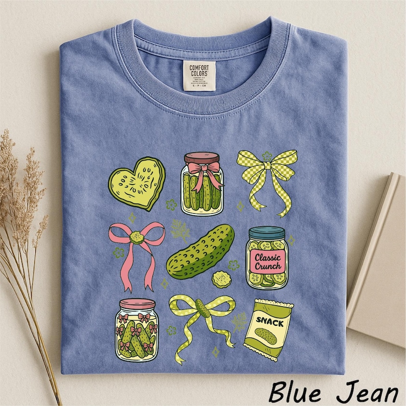 A blue t-shirt with a graphic design of various pickles and jars, along with a heart, bow, and other decorative elements.