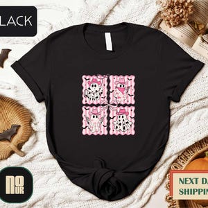 a black t-shirt with a graphic design of four cartoon-style cats wearing hats and scarves, arranged in a square pattern. The t-shirt is displayed on a bed with a blanket and a pumpkin, along with some bats and a book.