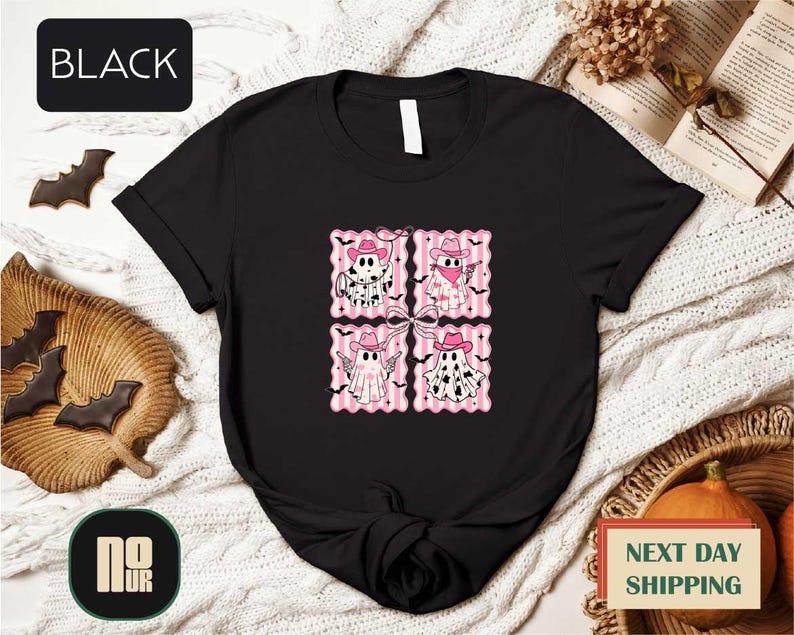 a black t-shirt with a graphic design of four cartoon-style cats wearing hats and scarves, arranged in a square pattern. The t-shirt is displayed on a bed with a blanket and a pumpkin, along with some bats and a book.