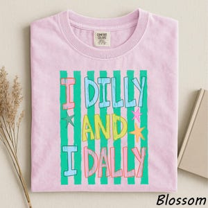A pink t-shirt with a colorful graphic design featuring the text "I Dilly and I Dally" in a playful font.