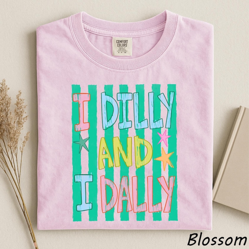 A pink t-shirt with a colorful graphic design featuring the text "I Dilly and I Dally" in a playful font.