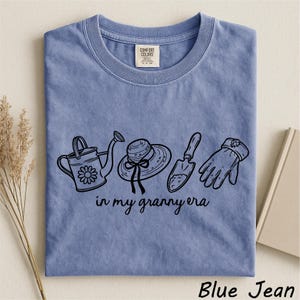 A blue t-shirt with a graphic design of gardening tools and accessories, including a watering can, a hat, and a shovel, along with the text "in my granny era" printed on it.