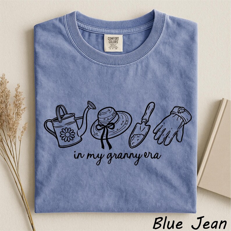 A blue t-shirt with a graphic design of gardening tools and accessories, including a watering can, a hat, and a shovel, along with the text "in my granny era" printed on it.