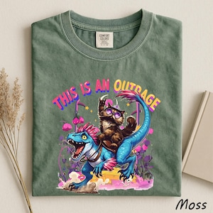 May include: Moss green t-shirt with a graphic of a cat riding a blue dinosaur, with the text "THIS IS AN OUTRAGE" in pink and yellow. The shirt is made of a soft material and has a crew neck.
