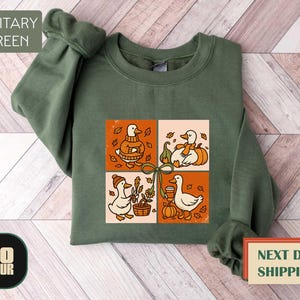 May include: Military green sweatshirt with a four-panel graphic featuring cartoon ducks in fall settings. Each panel shows a duck with pumpkins, leaves, and a basket. The sweatshirt has a crew neck and long sleeves.