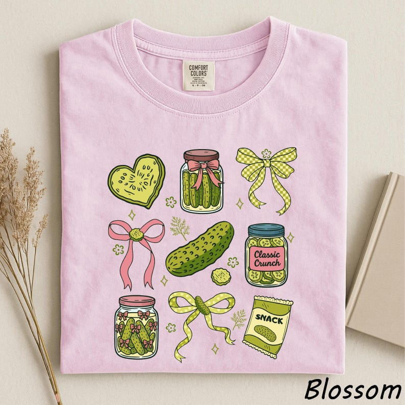 A pink t-shirt with various illustrations of pickles, jars, and other food items, along with a heart-shaped pickle and a classic crunch snack.