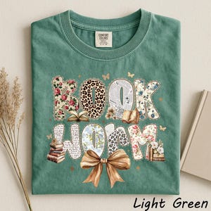 May include: Light green t-shirt with the words "BOOK WORM" in a decorative design. The letters are filled with floral, leopard print, and newspaper patterns. A brown bow is at the bottom. The shirt has a crew neck and short sleeves.