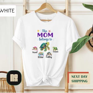May include: White t-shirt with the text "This MOM belongs to" and four sea turtles with names "Hazel", "River", "Oakley", and "Ocean" printed on it.