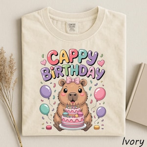 May include: Ivory-colored t-shirt featuring a cartoon capybara holding a birthday cake with candles. The shirt has the words "Cappy Birthday" in colorful bubble letters, with balloons and confetti. The shirt is made of a soft material.