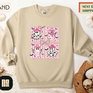 a beige sweatshirt with a graphic design of four pink ghost characters wearing cowboy hats and holding bats. The sweatshirt is displayed on a white background, and there is a wooden hat and a basket of pumpkins nearby.