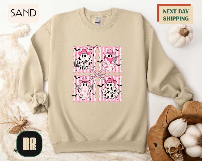 a beige sweatshirt with a graphic design of four pink ghost characters wearing cowboy hats and holding bats. The sweatshirt is displayed on a white background, and there is a wooden hat and a basket of pumpkins nearby.