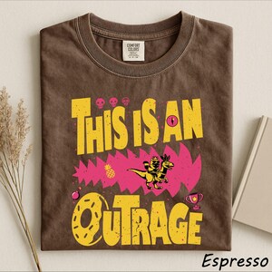 May include: A brown crew neck t-shirt featuring the yellow text THIS IS AN OUTRAGE. The graphic includes a pink jagged shape, a small dinosaur with a rider, three skulls, a pineapple, a donut, a trophy, and a Comfort Colors brand label.