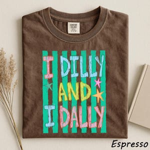 A brown t-shirt with a colorful graphic design featuring the text "I Dilly and I Dally" in a playful font.