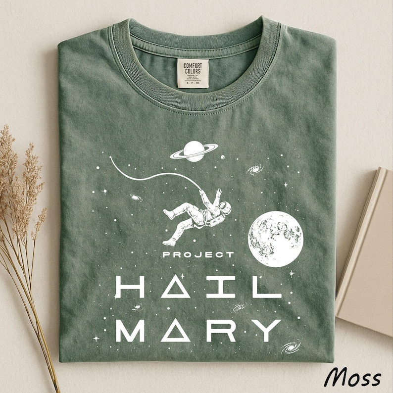 May include: A moss green crew neck t-shirt featuring a white graphic print of an astronaut floating in space with a tether, a planet with rings, and a moon. The text "PROJECT HAIL MARY" is printed in white below the astronaut in a modern font.