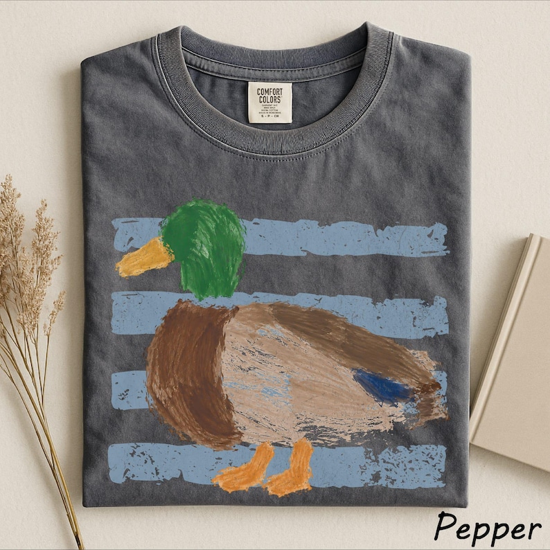 A gray t-shirt with a colorful drawing of a duck printed on it, along with some dried flowers and a book.