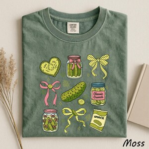 A green t-shirt with a variety of colorful illustrations, including jars of pickles, a heart-shaped object, a bow, and a bag of chips, all set against a light background.