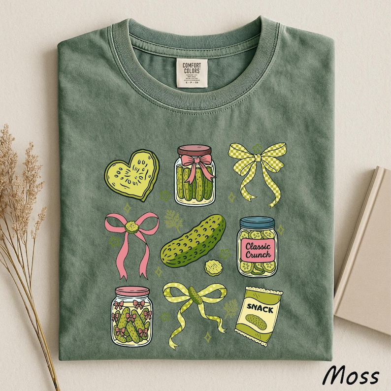 A green t-shirt with a variety of colorful illustrations, including jars of pickles, a heart-shaped object, a bow, and a bag of chips, all set against a light background.