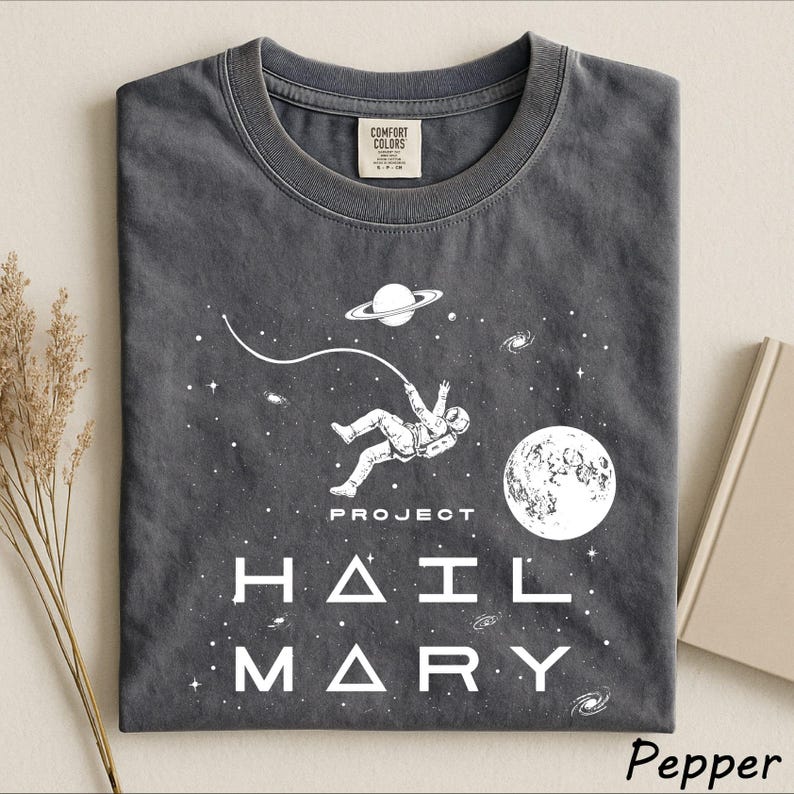 May include: A charcoal gray t-shirt featuring a white graphic of an astronaut floating in space with a tether, a planet with rings, a moon, and stars. The text reads PROJECT HAIL MARY in a stylized font. The shirt has a Comfort Colors brand label.