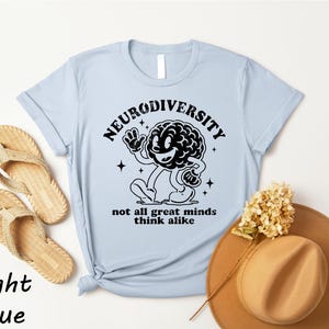 a light blue t-shirt with a graphic design of a brain and the text "Neurodiversity not all great minds think alike" printed on it. The t-shirt is placed on a white background, along with a pair of beige flip-flops, a brown hat, and some dried flowers.