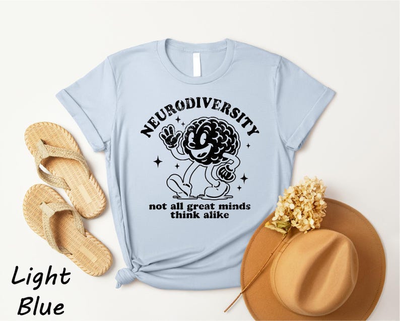 a light blue t-shirt with a graphic design of a brain and the text "Neurodiversity not all great minds think alike" printed on it. The t-shirt is placed on a white background, along with a pair of beige flip-flops, a brown hat, and some dried flowers.