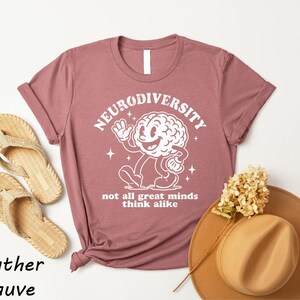 a pink t-shirt with a white graphic of a brain and the text "Neurodiversity" and "not all great minds think alike" printed on it, along with a pair of beige flip-flops and a beige hat.