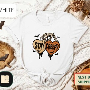 a white t-shirt with a graphic design that says "STAY CREEPY" in black text, surrounded by bats and a skeleton hand. The t-shirt is displayed on a white background, with other Halloween-themed items such as bats and a pumpkin visible in the background.