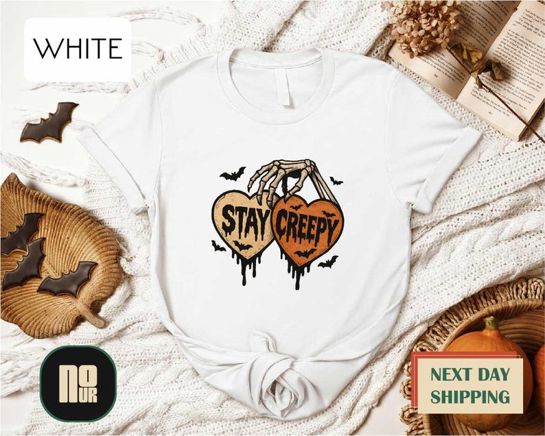 a white t-shirt with a graphic design that says "STAY CREEPY" in black text, surrounded by bats and a skeleton hand. The t-shirt is displayed on a white background, with other Halloween-themed items such as bats and a pumpkin visible in the background.
