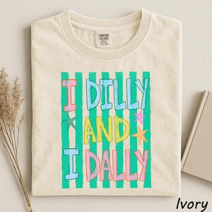 A beige t-shirt with a colorful graphic design featuring the text "I Dilly and I Dally" in a playful font.