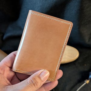 May include: A tan leather wallet with a polished surface. The edges are stitched with a pale thread. The wallet is rectangular and appears unused.