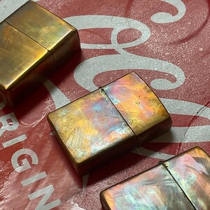 Pode incluir: Tr&ecirc;s isqueiros de lat&atilde;o vintage com um acabamento iridescente desgastado. Os isqueiros s&atilde;o retangulares com uma tampa articulada. O fundo &eacute; uma superf&iacute;cie vermelha com letras brancas.