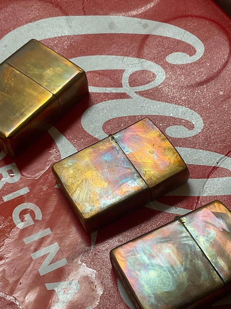 Pode incluir: Tr&ecirc;s isqueiros de lat&atilde;o vintage com um acabamento iridescente desgastado. Os isqueiros s&atilde;o retangulares com uma tampa articulada. O fundo &eacute; uma superf&iacute;cie vermelha com letras brancas.