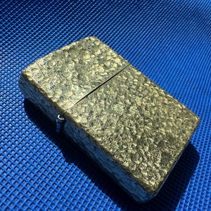 May include: A rectangular metal lighter with a gold-toned finish and a textured, hammered surface. The lighter features a hinged lid design with a visible metal hinge on the side, showcasing a reflective, uneven pattern across the entire exterior.