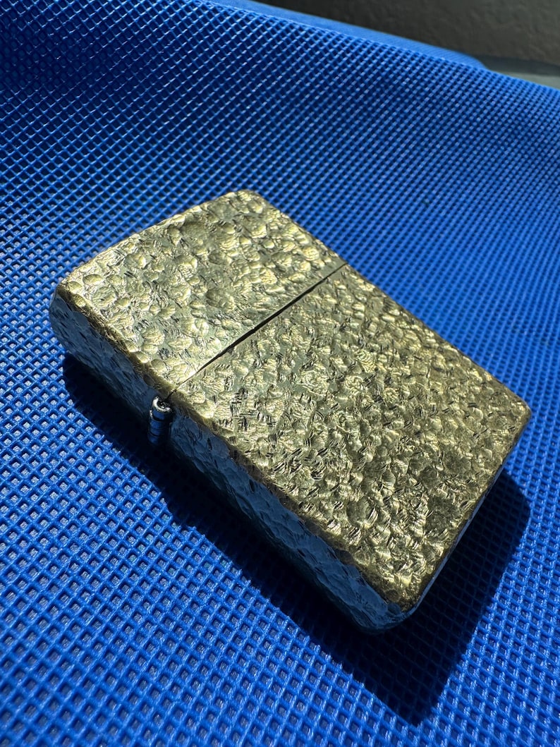 May include: A rectangular metal lighter with a gold-toned finish and a textured, hammered surface. The lighter features a hinged lid design with a visible metal hinge on the side, showcasing a reflective, uneven pattern across the entire exterior.