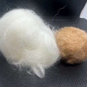 May include: A large, fluffy, white ball of raw wool fiber positioned next to a smaller, denser, tan-colored ball of felted wool. Both objects exhibit a soft, fibrous texture with loose strands visible on the surface of the white wool.