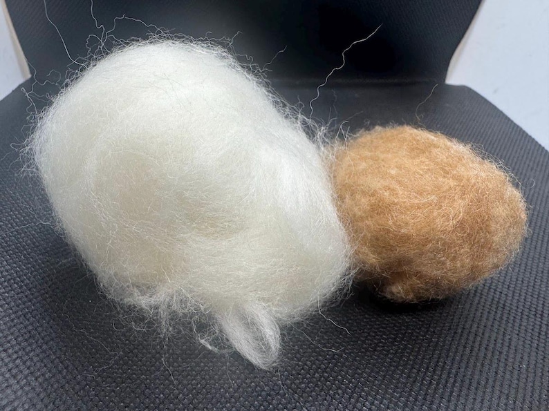 May include: A large, fluffy, white ball of raw wool fiber positioned next to a smaller, denser, tan-colored ball of felted wool. Both objects exhibit a soft, fibrous texture with loose strands visible on the surface of the white wool.