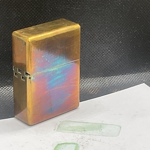 Pode incluir: Um isqueiro de lat&atilde;o vintage com formato retangular. O isqueiro tem um acabamento p&aacute;tina com tons iridescentes azuis e vermelhos. A dobradi&ccedil;a e o fecho s&atilde;o vis&iacute;veis na lateral. O isqueiro est&aacute; sobre uma superf&iacute;cie branca.