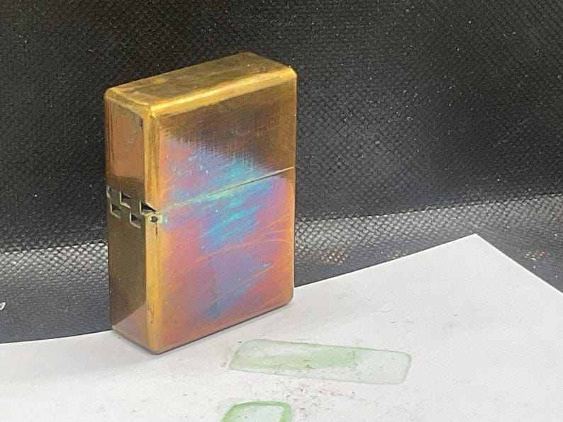 Pode incluir: Um isqueiro de lat&atilde;o vintage com formato retangular. O isqueiro tem um acabamento p&aacute;tina com tons iridescentes azuis e vermelhos. A dobradi&ccedil;a e o fecho s&atilde;o vis&iacute;veis na lateral. O isqueiro est&aacute; sobre uma superf&iacute;cie branca.