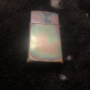 Pode incluir: Um isqueiro retangular iridescente com acabamento em metal escovado. A superf&iacute;cie do isqueiro exibe um espectro de cores, incluindo azuis, verdes e roxos. O isqueiro est&aacute; fechado e tem uma tampa articulada.