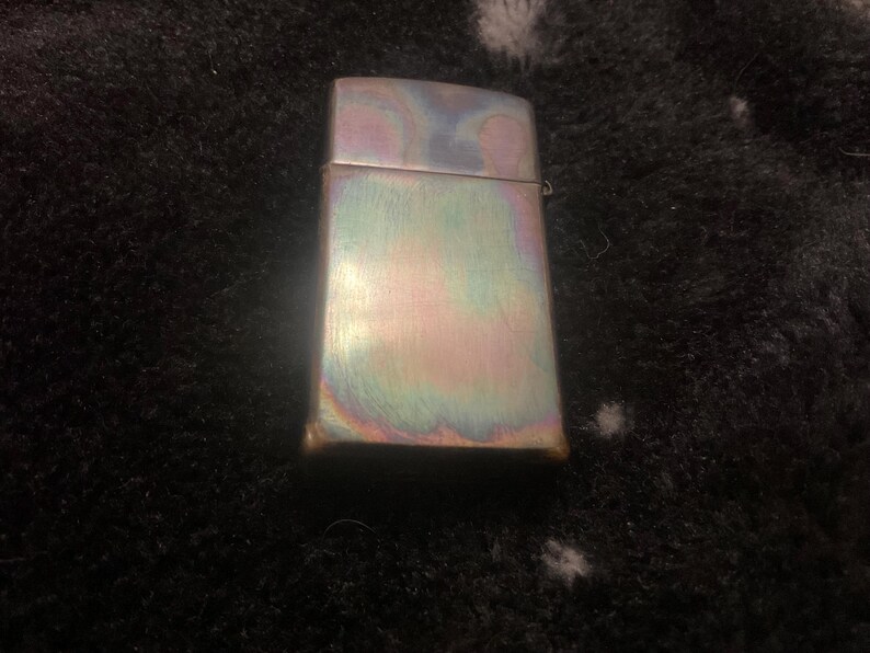 Pode incluir: Um isqueiro retangular iridescente com acabamento em metal escovado. A superf&iacute;cie do isqueiro exibe um espectro de cores, incluindo azuis, verdes e roxos. O isqueiro est&aacute; fechado e tem uma tampa articulada.