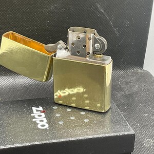 以下が含まれることがあります： 蓋が開いた状態の光沢のあるゴールドトーンの金属製ライター。銀色のチムニーとフリントホイール機構が見えています。ライターは、上面に白いZippoのロゴが印刷された黒い長方形の箱の上に置かれています。