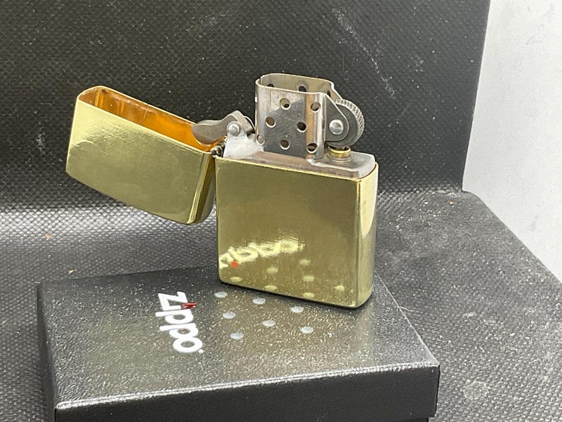 以下が含まれることがあります： 蓋が開いた状態の光沢のあるゴールドトーンの金属製ライター。銀色のチムニーとフリントホイール機構が見えています。ライターは、上面に白いZippoのロゴが印刷された黒い長方形の箱の上に置かれています。