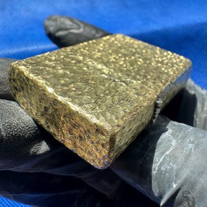May include: A rectangular gold-colored lighter featuring a heavily textured, hammered surface finish. The metal casing displays a reflective, uneven pattern across its top and side panels, with a visible hinge mechanism located on the right edge.