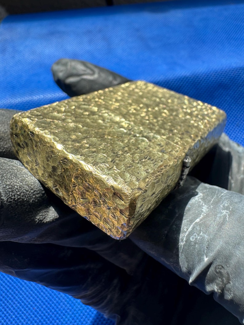 May include: A rectangular gold-colored lighter featuring a heavily textured, hammered surface finish. The metal casing displays a reflective, uneven pattern across its top and side panels, with a visible hinge mechanism located on the right edge.